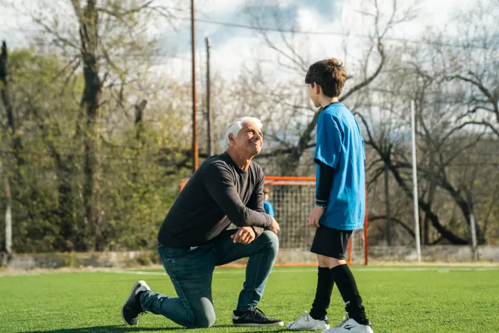 Un abuelo arrodillado en la cancha conversa con su nieto vestido con ropa de fútbol
